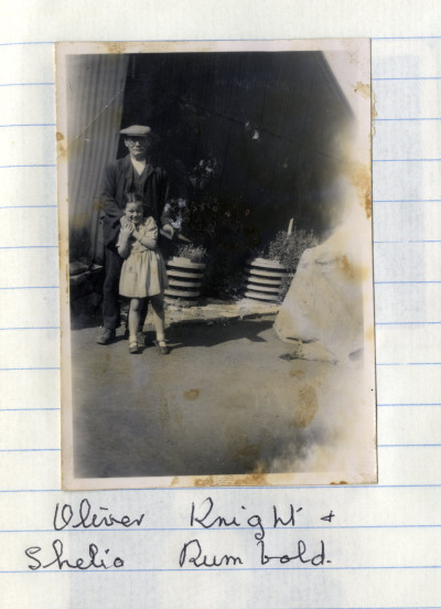 Older man in cloth cap holding small girl by withy shed